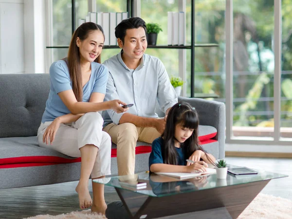 Millennial Asian happy family father and mother sitting on cozy sofa couch holding remote watching television while little young girl daughter sitting on floor doing homework in living room at home.