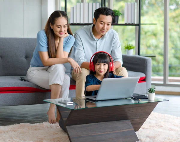 Millennial Asian happy family father and mother sit on cozy sofa couch smiling helping teaching little young girl daughter wearing headphones on floor studying learning online in living room at home.