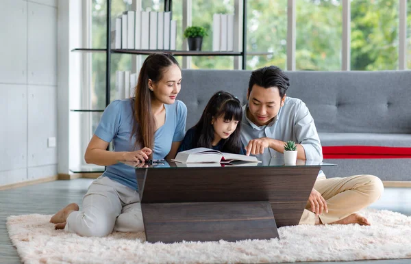 Millennial Asian happy family father and mother sitting on cozy carpet floor smiling helping teaching little young girl daughter reading studying learning with big textbook in living room at home.
