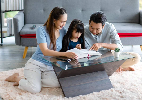 Millennial Asian happy family father and mother sitting on cozy carpet floor smiling helping teaching little young girl daughter reading studying learning with big textbook in living room at home.