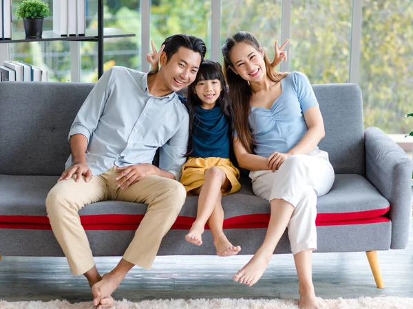 Millennial Asian happy family father mother and little young child girl sitting smiling looking at camera together on cozy sofa couch showing two fingers peace sign taking photo in living room home.