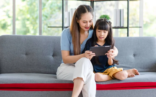 stock image Millennial Asian happy family mother sitting smiling on cozy sofa couch holding showing funny content streaming online via touchscreen smartphone to little young girl daughter in living room at home.