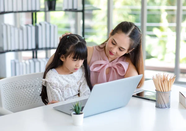 Millennial Asian happy family mother smiling helping supporting teaching little girl kid daughter studying learning doing online school homework via laptop notebook computer in living room at home.