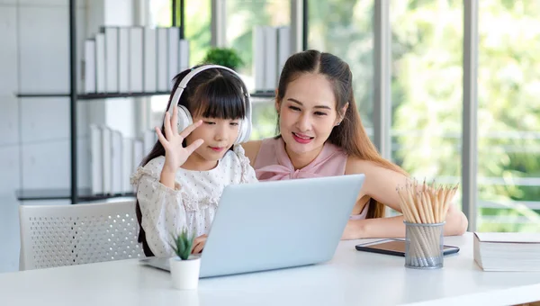 Millennial Asian happy family mother smiling helping supporting teaching little girl kid daughter studying learning doing online school homework via laptop notebook computer in living room at home.