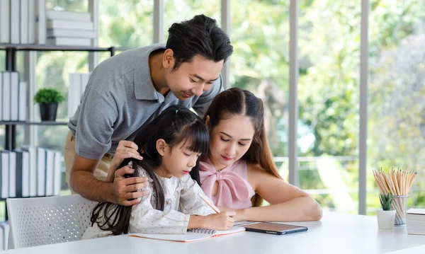 Millennial Asian happy family father and mother smiling helping supporting teaching little girl daughter studying learning writing doing school homework via tablet computer in living room at home.