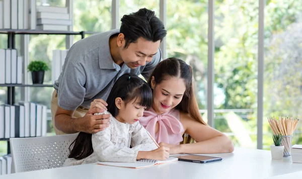 Millennial Asian happy family father and mother smiling helping supporting teaching little girl daughter studying learning writing doing school homework via tablet computer in living room at home.