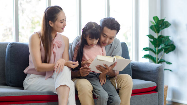 Тысячелетняя азиатская счастливая семья мать отец сидит на уютном диване, улыбаясь вместе обучая маленькая девочка дочь изучает чтение художественный роман учебник в гостиной на дому.