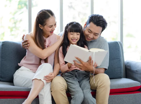 Millennial Asian happy family father mother sitting on cozy sofa couch smiling together teaching little girl kid daughter learning studying reading fiction novel textbook in living room at home.