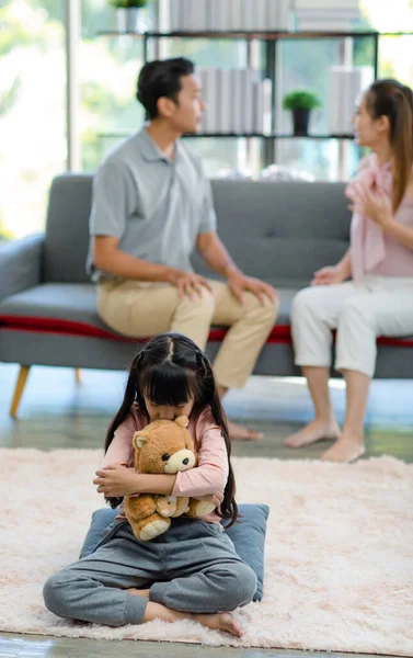 Asian unhappy sad depressed stressed little girl daughter sitting on carpet floor hugging holding teddy bear doll crying alone while father and mother fighting having argument in living room at home.