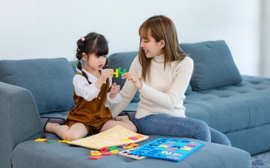Millennial Asian happy cheerful little cute preschooler daughter girl playing learning alphabets letters jigsaw toy on sofa in living room while young teenager mother nanny helping teaching supporting