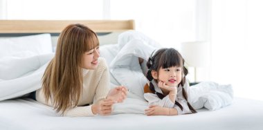 Millennial Asian young beautiful mother in sweater waking up laying down smiling together under thick warm blanket on bed in morning with little cute preschooler daughter girl in bedroom at home.