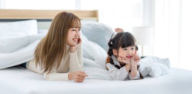 Millennial Asian young beautiful mother in sweater waking up laying down smiling together under thick warm blanket on bed in morning with little cute preschooler daughter girl in bedroom at home.