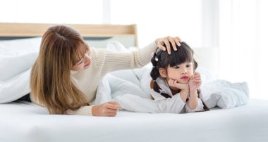 Millennial Asian young beautiful mother in sweater waking up laying down smiling together under thick warm blanket on bed in morning with little cute preschooler daughter girl in bedroom at home.