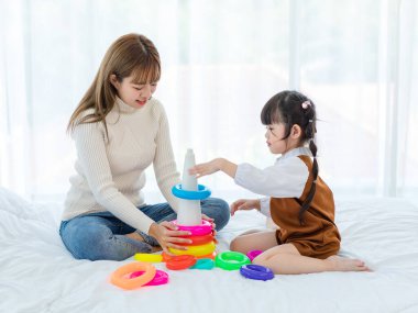 Milenyum Asyalı genç, güzel, gündelik kıyafetli genç bir anne kucağında gülücüklerle eğlenceli lastik yüzükler oynuyor küçük tatlı anaokulu kızıyla oynayan çocuklar..