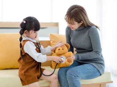 Milenyum Asyalı genç bayan anne rahat koltukta oturmuş gülümsüyor oyuncak ayı bebeği tutuyor ve steteskoplu küçük tatlı bir anaokulu kızıyla doktorculuk oynuyor..