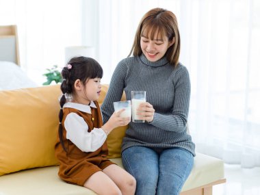 Millennial Asian young pretty female teenager mother nanny babysitter in casual outfit sitting on sofa smiling holding serving delicious milk glass to little cute preschooler daughter girl drinking.
