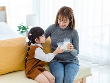 Millennial Asian young pretty female teenager mother nanny babysitter in casual outfit sitting on sofa smiling holding serving delicious milk glass to little cute preschooler daughter girl drinking.