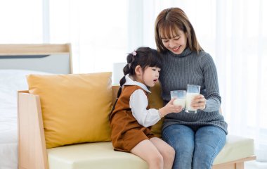 Millennial Asian young pretty female teenager mother nanny babysitter in casual outfit sitting on sofa smiling holding serving delicious milk glass to little cute preschooler daughter girl drinking.