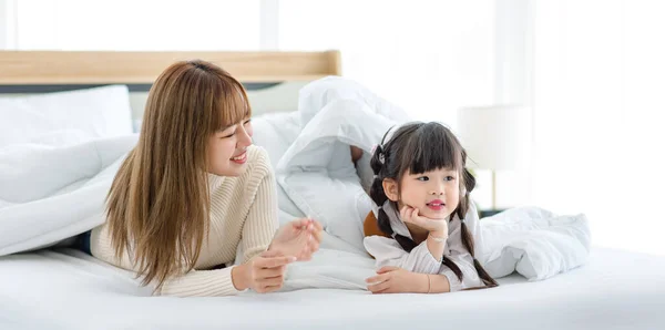 stock image Millennial Asian young beautiful mother in sweater waking up laying down smiling together under thick warm blanket on bed in morning with little cute preschooler daughter girl in bedroom at home.