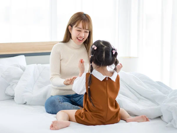 stock image Millennial Asian young female teenager mother nanny babysitter in casual sweater and jeans sitting playing with little cute preschooler daughter girl on bed in bedroom.