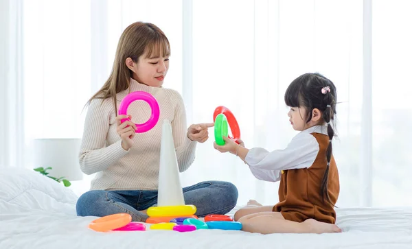 千年亚洲年轻漂亮的少女妈妈穿着休闲装坐在床上笑着拿着玩具娃娃玩着创意的橡皮圈和可爱的小女孩玩着玩具 — 图库照片