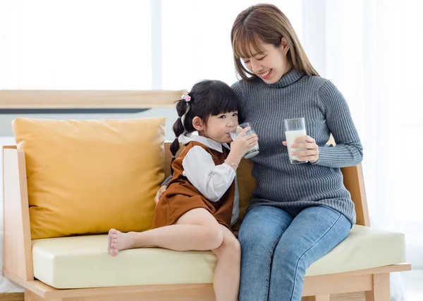 Millennial Asian young pretty female teenager mother nanny babysitter in casual outfit sitting on sofa smiling holding serving delicious milk glass to little cute preschooler daughter girl drinking.