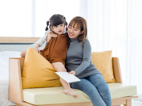 Millennial Asian cheerful happy young pretty female teenager mother nanny babysitter in casual outfit sitting on sofa smiling hugging little cute preschooler daughter girl after receive lovely card.