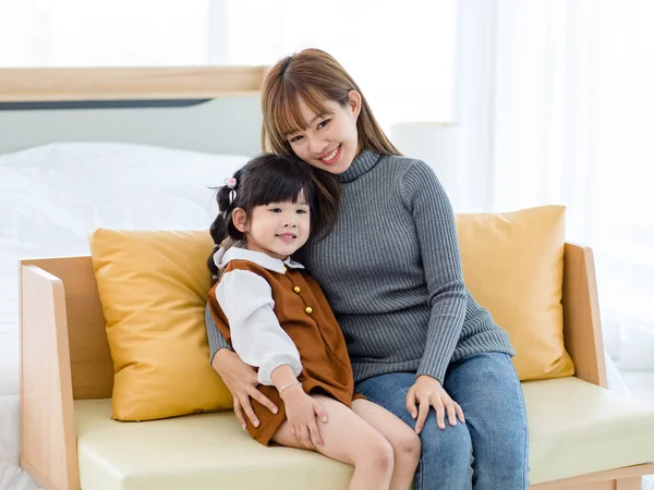 Millennial Asian cheerful happy young pretty female teenager mother nanny babysitter in casual outfit sitting on cozy sofa smiling hugging showing love with little cute preschooler daughter girl.