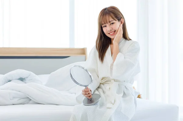 Millennial Asian cheerful happy young beautiful female teenager housewife in white bathrobe sitting smiling holding hand touching cheek looking at mirror on bed in bedroom in morning after waking up.