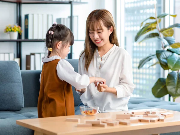 Millennial Asian cheerful happy young beautiful female teenager mother nanny babysitter sitting smiling holding delicious jelly hiding from little cute preschooler daughter girl at home living room.