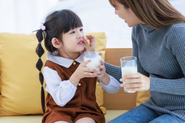 Millennial Asian young pretty female teenager mother nanny babysitter in casual outfit sitting on sofa smiling holding serving delicious milk glass to little cute preschooler daughter girl drinking.