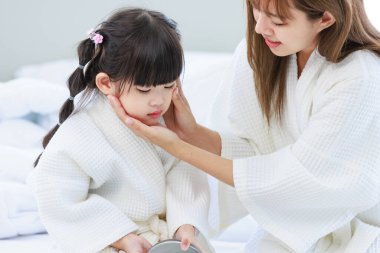 Millennial Asyalı mutlu, neşeli, küçük tatlı anaokulu kızı banyodan sonra beyaz bornoz giymiş el aynasına bakıyor tanınmayan bir anneyle saç stili yapıyor..