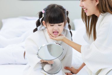 Millennial Asyalı mutlu, neşeli, küçük tatlı anaokulu kızı banyodan sonra beyaz bornoz giymiş el aynasına bakıyor tanınmayan bir anneyle saç stili yapıyor..