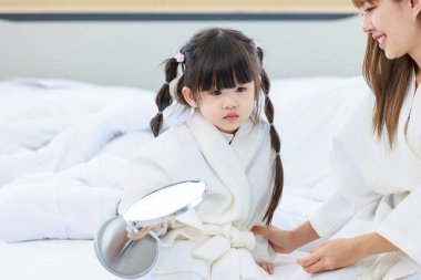 Millennial Asyalı mutlu, neşeli, küçük tatlı anaokulu kızı banyodan sonra beyaz bornoz giymiş el aynasına bakıyor tanınmayan bir anneyle saç stili yapıyor..