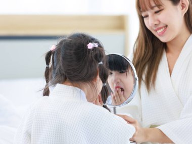 Millennial Asyalı mutlu, neşeli, küçük tatlı anaokulu kızı banyodan sonra beyaz bornoz giymiş el aynasına bakıyor tanınmayan bir anneyle saç stili yapıyor..