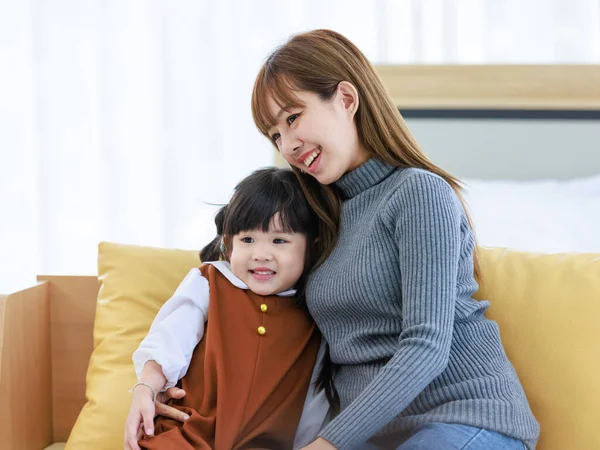 Millennial Asian cheerful happy young pretty female teenager mother nanny babysitter in casual outfit sitting on cozy sofa smiling hugging showing love with little cute preschooler daughter girl.