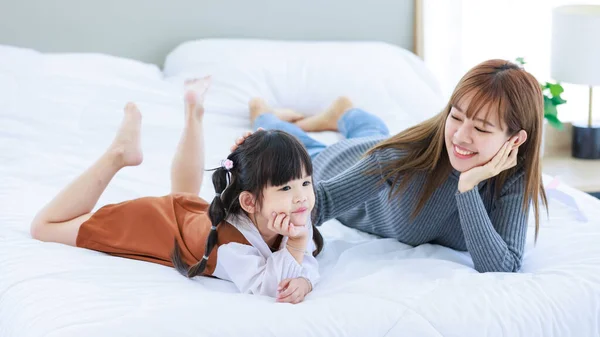 Millennial Asian young pretty female teenager mother nanny babysitter in casual outfit laying down on bed smiling thinking with little cute preschooler thoughtful daughter girl in bedroom in morning.