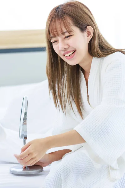 Millennial Asian cheerful happy young beautiful female teenager housewife in white bathrobe sitting smiling holding hand touching cheek looking at mirror on bed in bedroom in morning after waking up.