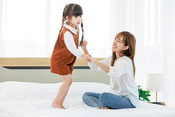 Millennial Asian happy cheerful little cute preschooler daughter girl standing dancing jumping on bed sheet in bedroom at home while young teenager mother nanny babysitter smiling helping supporting.