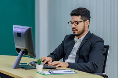 Milenyum Asyalı profesyonel Hintli sakallı iş adamı resmi takım elbiseli ve gözlüklü adam çok uluslu şirket ofisindeki çalışma masasında bilgisayar klavyesiyle çalışıyor..