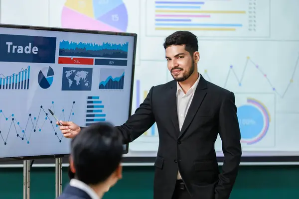 Millennial Asian Indian professional successful bearded male businessman presenter speaker in formal business suit hold pen point trading buy sell presentation monitor present to audiences in office.