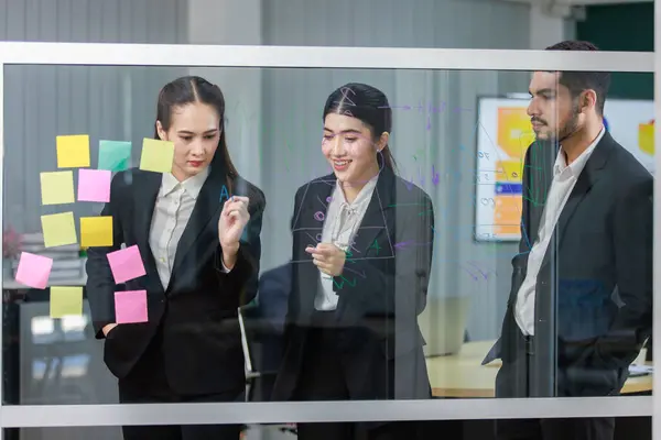 Millennial Asian professional successful female businesswoman in formal business suit standing using marker writing ideas strategy on glass board meeting with multinational colleagues in meeting room.