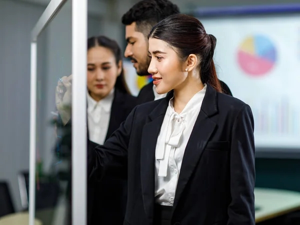 Millennial Asian professional successful female businesswoman in formal business suit standing using marker writing ideas strategy on glass board meeting with multinational colleagues in meeting room.