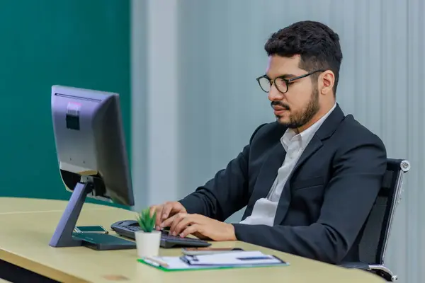 Millennial Asian Indian professional successful bearded male businessman in formal business suit and eyeglasses typing working with computer keyboard on work desk in multinational company office.
