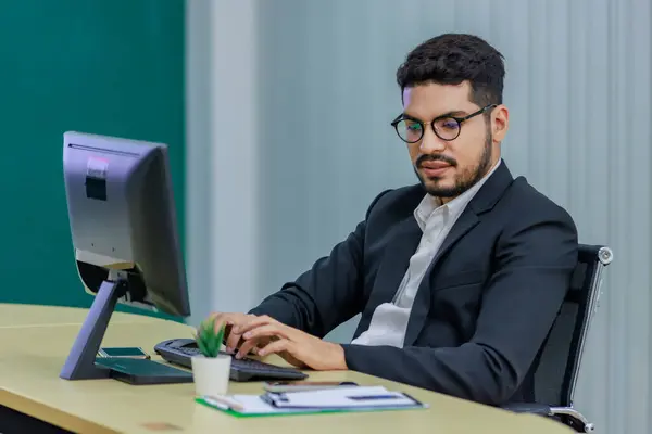 Millennial Asian Indian professional successful bearded male businessman in formal business suit and eyeglasses typing working with computer keyboard on work desk in multinational company office.