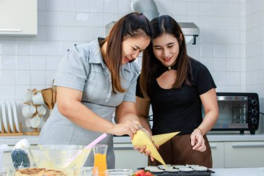 Asyalı mutlu kadın pastacı fırıncı aşçı ev hanımı. Yakın arkadaşları gülümseyerek ayakta duruyor. Tüp torba krem sıkarak, krem şanti sıkarak mutfakta kek hazırlıyor..