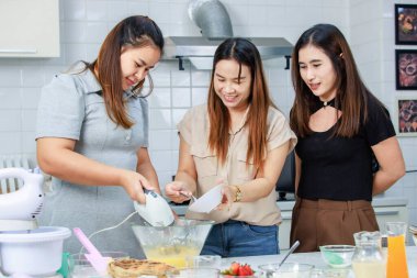 Asyalı mutlu kadın pastacı fırıncı aşçı ev hanımı. Yakın arkadaşları gülümseyerek yumurta çırpma makinesi kullanmalarına yardım ediyor. Süslü mutfakta hamur hazırlıyor..