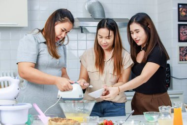 Asyalı mutlu kadın pastacı fırıncı aşçı ev hanımı. Yakın arkadaşları gülümseyerek yumurta çırpma makinesi kullanmalarına yardım ediyor. Süslü mutfakta hamur hazırlıyor..