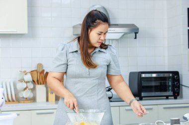 Milenyum Asyalı profesyonel kadın fırın şefi ev hanımı cam kâseye yumurta kırıyor çırpıyor un çırpıyor un hamuru pişiriyor tam donanımlı mutfakta lezzetli bir tatlı pişiriyor..