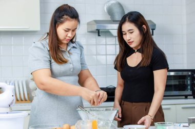 Asyalı profesyonel kadın pastane şefi ev hanımı ve arkadaşı sütçülerin taze sütü, dekore edilmiş mutfakta cam bardağa doldurup lezzetli bir tatlı pişirmeye yardım ediyorlar..